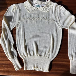 May include: A cream-colored long-sleeve knit sweater featuring a delicate crocheted Peter Pan collar, puffed shoulders, and a decorative diamond-patterned stitch across the chest. The garment includes a ribbed hem and cuffs.