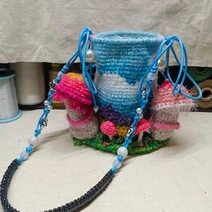 May include: A cylindrical crochet bag featuring a blue and white sky pattern, adorned with three-dimensional crocheted mushrooms in pink and beige. The bag includes a blue cord strap decorated with white beads and a black beaded section.