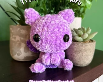 Puede incluir: Un juguete de peluche tejido a ganchillo de color morado con grandes ojos negros de plástico y orejas puntiagudas. El texto "mewtwo plush" aparece arriba y "PigeonPatternCrochet" abajo. El peluche tiene una textura suave y esponjosa.