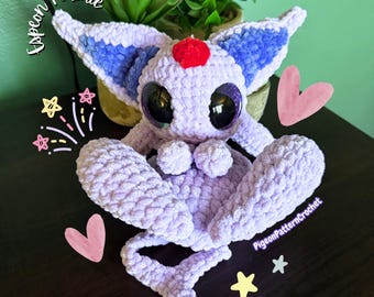 Puede incluir: Un peluche de ganchillo de color violeta claro con grandes ojos negros, orejas interiores de color violeta oscuro y una gema roja en la frente. El peluche tiene una cola larga y está decorado con gráficos digitales que incluyen el texto Espeon plushie y PigeonPatternCrochet.