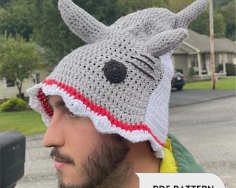 Puede incluir: Un gorro de tiburón tejido a ganchillo en color gris con un borde festoneado blanco y rojo, dos aletas dorsales puntiagudas en la parte superior y un ojo circular negro. Incluye un panel trasero blanco y un globo con el texto PDF PATTERN.