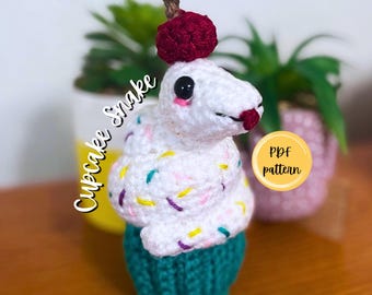 Puede incluir: Una figura de serpiente tejida a ganchillo en color blanco con chispas de colores, una cereza roja oscura en la cabeza y una base acanalada color verde azulado. El texto "Cupcake Snake" y "PDF pattern" son visibles, junto a "PigeonPatternCrochet".