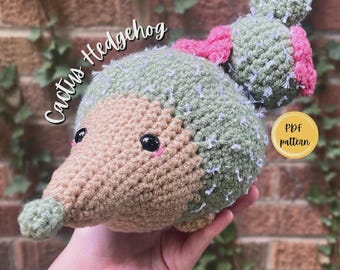 Puede incluir: Un juguete de erizo tejido a ganchillo con cara color canela, cuerpo verde salvia con detalles blancos texturizados y una pequeña nariz verde. La espalda incluye una estructura de cactus verde con flores rosas. Texto Cactus Hedgehog y PDF pattern.