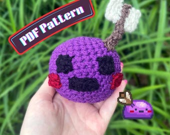 Puede incluir: Una esfera de ganchillo morada con ojos y boca negros, mejillas rojas y un hacha blanca unida a la parte superior. La imagen incluye el texto PDF Pattern y AbiesCustomDesigns, junto con un pequeño gráfico pixelado de una criatura morada.