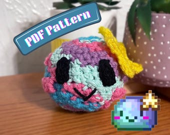 Puede incluir: Un juguete de peluche tejido a ganchillo de forma redonda con un patrón de hilo jaspeado en tonos rosa, azul claro y morado. Tiene dos ojos ovalados negros, una boca cosida negra y una estrella tejida amarilla en el lateral. Texto: PDF Pattern.