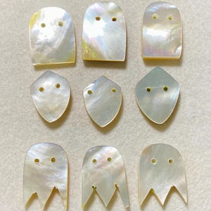 May include: Nine iridescent mother-of-pearl buttons arranged in three rows. The top row features three rounded rectangular buttons, the middle row has three shield-shaped buttons, and the bottom row displays three ghost-shaped buttons with two holes.