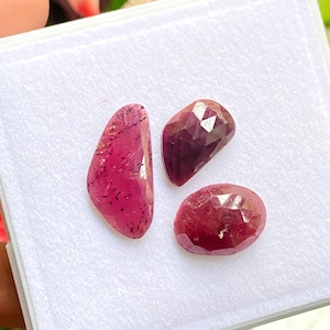 Puede incluir: Tres rubíes facetados sueltos sobre una base de espuma blanca. Una piedra es alargada y tiene forma de lágrima con inclusiones oscuras, mientras que las otras dos son más pequeñas, facetadas y de color rojo intenso, con forma de pera y ovalada.