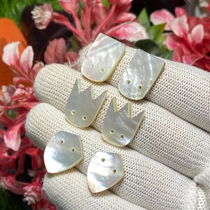 May include: Six iridescent mother-of-pearl buttons arranged in three pairs on a textured white fabric surface. The top pair is rectangular with rounded corners, the middle pair is tulip-shaped, and the bottom pair is pentagonal, each with two holes.