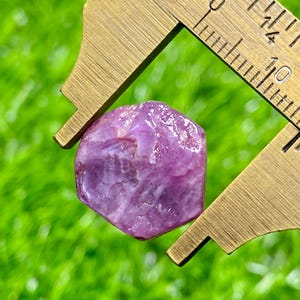 May include: A raw, hexagonal-shaped purple gemstone held within the jaws of a brass caliper. The metal tool features clear metric markings, showing the stone measures approximately 6 millimetres in width across its widest point.