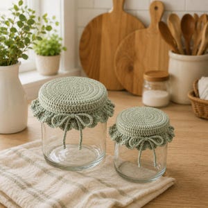 Può includere: Due barattoli in vetro trasparente con coperture all'uncinetto color verde salvia. Ogni coperchio presenta una parte superiore circolare lavorata all'uncinetto, un bordo arricciato e un piccolo fiocco decorativo legato sul davanti.