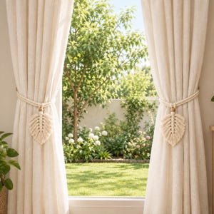 May include: A pair of off-white linen-textured curtains held back by two macrame tiebacks. Each tieback features a braided cord, a single round wooden bead, and a decorative leaf-shaped macrame tassel in a matching off-white cotton cord.