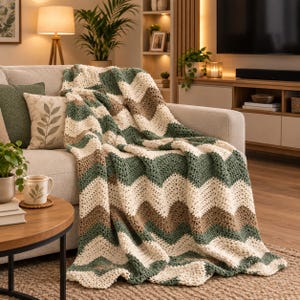 Peut inclure: Un jeté de canapé crocheté présentant un motif à chevrons répétitif avec des rayures alternées vert sauge, crème et marron clair. Le textile possède une construction en mailles texturées et est drapé sur l'assise d'un canapé clair.