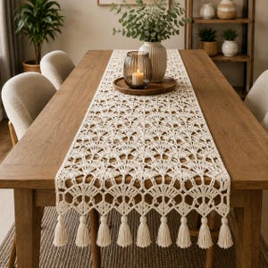Puede incluir: Un camino de mesa de ganchillo color crema con un intrincado patrón en forma de abanico y un borde festoneado acabado con largas borlas colgantes. El camino se extiende plano sobre una mesa de madera, mostrando su detallado diseño calado.