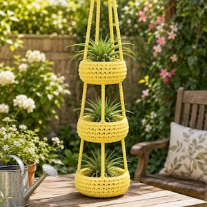 Puede incluir: Un soporte para plantas colgante de tres niveles hecho de hilo de ganchillo amarillo brillante. Cada una de las tres cestas circulares contiene una planta de aire verde puntiaguda. La estructura se suspende mediante cordones de hilo trenzado.