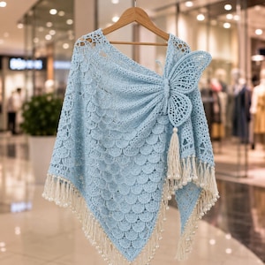 May include: A light blue crochet poncho featuring a prominent butterfly motif on the shoulder, a scalloped stitch pattern throughout the body, and a bottom hem finished with cream-colored fringe tassels accented with small white beads.