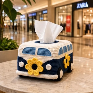 May include: A crochet tissue box cover shaped like a van in white and dark blue. The van features light blue windows, black and white wheels, and large yellow flower appliqués with dark centers on the front and side panels. White tissues emerge.