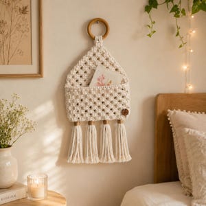 May include: A cream-colored macrame wall hanging featuring a triangular pocket design suspended from a circular wooden ring. The piece includes four long, thick tassels at the bottom and a small circular wooden tag on the lower right side.