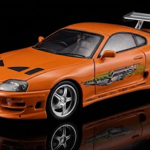 May include: A die-cast model of an orange sports car featuring a large silver rear spoiler, silver five-spoke wheels, and green graphic decals along the side panels. The vehicle has a front bumper with air intakes and black hood vents.