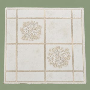 May include: A square ceramic tile featuring a grid pattern outlined in light brown lines. The tile has a textured off-white surface with two diagonal squares decorated with a light brown floral motif consisting of small blossoms and leaves.