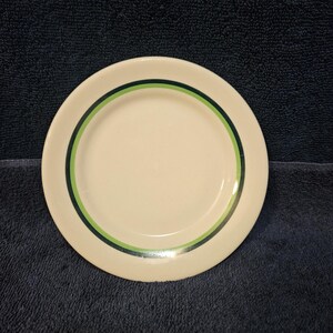 May include: A round, off-white ceramic plate featuring a decorative border consisting of a thin dark green ring encircling a wider light green band, creating a simple and clean design on the surface of the dishware.