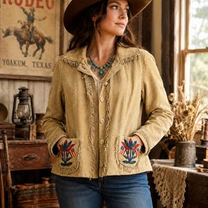 May include: A tan suede jacket featuring a collared neckline, long sleeves, and a front button closure. The jacket is adorned with fringe trim along the lapels and pockets, and includes two front pockets with embroidered floral and flag motifs.