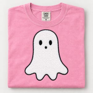 以下が含まれることがあります： ピンク色のクルーネックTシャツ。黒い刺繍の目と小さな口を持つ、白いテクスチャーのあるシェニール織のゴーストの大きなワッペンが付いています。内側のラベルには「Comfort Colors」、100%リングスパンコットン、サイズMと記載されています。