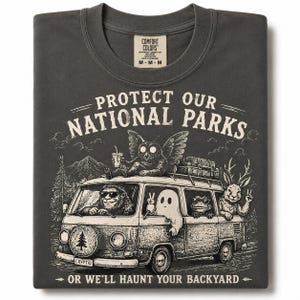 May include: A charcoal gray t-shirt featuring a graphic of a van with a Bigfoot driver, a ghost, a frog, and a jackalope passenger. An owl sits on the roof. The text reads: PROTECT OUR NATIONAL PARKS OR WE'LL HAUNT YOUR BACKYARD. Comfort Colors label.
