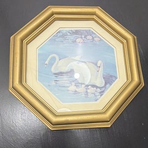 May include: An octagonal gold-toned frame surrounds a print depicting two white adult swans and three small cygnets swimming in a blue pond. The scene includes water lilies and lily pads in the background, all protected by a glass cover.