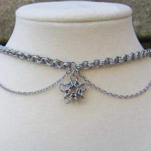 May include: A silver-toned chainmail choker necklace featuring a central star-shaped charm constructed from interlocking metal rings. The design includes a thicker woven chainmail band with two thinner, draping silver chains attached.