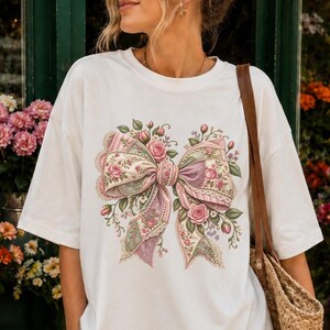 Puede incluir: Una camiseta blanca de manga corta con un gran gráfico central de un lazo decorativo adornado con intrincados patrones florales en tonos rosa, verde y morado. La camiseta tiene un cuello redondo y un corte holgado, combinada con pantalones cortos.