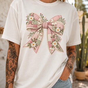 May include: A white short-sleeved t-shirt featuring a large central graphic of a bow. The bow is composed of various patchwork patterns in shades of pink, white, and sage green, decorated with embroidered white daisies and small pink roses.