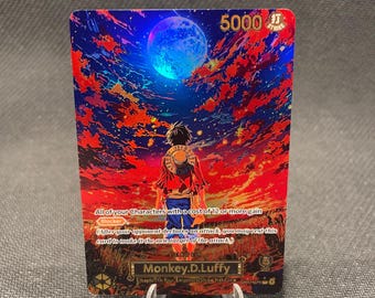 Puede incluir: Una tarjeta coleccionable rectangular con una ilustración vibrante de un personaje con sombrero de paja de pie en un campo bajo una luna azul y un cielo rojo, naranja y morado. La tarjeta muestra el texto Monkey D. Luffy y estadísticas de juego.