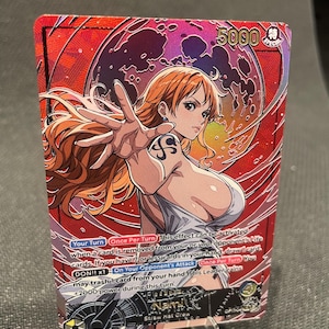 May include: A One Piece trading card featuring Nami with orange hair and a shoulder tattoo, set against a red background with a dark moon. The card displays the text Nami, Straw Hat Crew, 5000 power, and game instructions in English text.