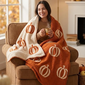 May include: A reversible plush throw blanket featuring a pumpkin pattern. One side is cream-coloured with burnt orange pumpkins, and the other side is burnt orange with cream-coloured pumpkins. The fabric has a textured, fuzzy, fleece-like finish.