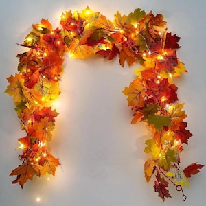 Puede incluir: Una guirnalda decorativa con hojas de arce artificiales en tonos rojo, naranja, amarillo y verde. El follaje está entrelazado con finos hilos de alambre flexibles que contienen pequeñas luces LED de color blanco cálido que emiten un brillo.