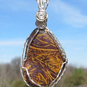 May include: A teardrop-shaped pendant with a silver wire wrap and chain. The pendant features a polished stone with a maroon base and yellow-gold linear patterns. The background is a blurred view of a blue sky and foliage.