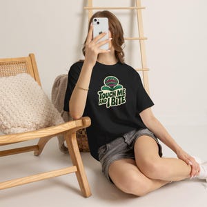May include: A black t-shirt featuring a graphic of a green carnivorous plant with sharp teeth and the text "TOUCH ME AND I BITE" in cream-coloured lettering. The design is centred on the chest of the shirt, which has short sleeves and a crew neck.