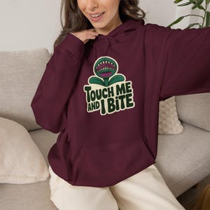 May include: A maroon hooded sweatshirt featuring a graphic of a green carnivorous plant with sharp teeth. The text "TOUCH ME AND I BITE" is printed in dark green letters with a cream-colored outline below the plant illustration on the chest.
