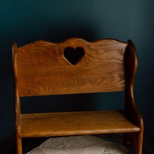 May include: A small wooden bench with a medium brown finish and a visible wood grain. The backrest features a scalloped top edge and a centred heart-shaped cutout. The bench has two side panels and a flat seat, constructed from solid wood.