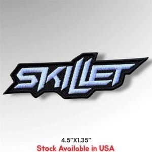 May include: An embroidered patch featuring the word SKILLET in a stylized, angular font with light blue lettering outlined in black. The patch measures 4.5 inches by 1.35 inches and has a textured, fabric-like appearance with visible stitching.