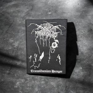 May include: A rectangular black fabric patch featuring a white screen-printed graphic. The design includes stylized, jagged lettering at the top, a central figure with an open mouth, and the text Transilvanian Hunger printed in a gothic font at the base.