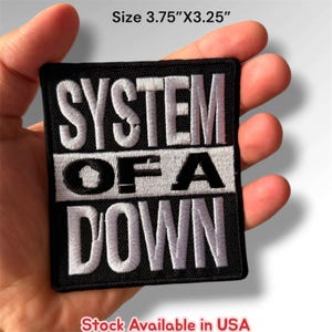 May include: A rectangular embroidered patch featuring the text SYSTEM OF A DOWN in white and black thread on a black background. The patch measures 3.75 inches by 3.25 inches and has a stitched border. The design consists of three horizontal rows.