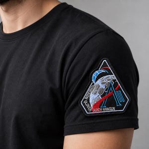 May include: A black short-sleeved t-shirt featuring a triangular embroidered patch on the left sleeve. The patch depicts a moon surface, a blue letter A, and red orbital lines, with the text WISEMAN, GLOVER, and KOCH HANSEN stitched in white.