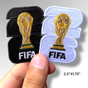 May include: Two embroidered patches measuring 2.5 inches by 1.75 inches. One patch is black and the other is white, both featuring a gold-colored FIFA World Cup trophy graphic and the text FIFA in bold, block lettering at the bottom.