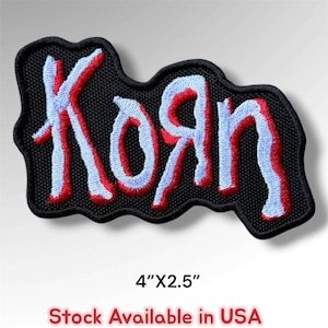 May include: An embroidered patch featuring the band name Korn in a stylized, jagged font. The lettering is light blue with a red outline, set against a solid black background. The patch measures 4 inches by 2.5 inches as indicated by text.
