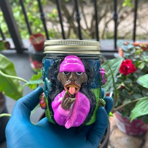 Könnte beinhalten: Ein Glasgefäß mit einer dreidimensionalen skulptierten Figur, die eine pinke Mütze, eine pinke Jacke und ein goldenes Grill trägt, die Zunge herausstreckt und eine Handgeste macht. Das Glas hat einen metallischen Schraubdeckel und einen dunkelgrünen und blauen Hintergrund.