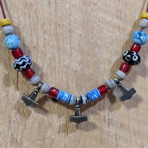 Peut inclure: Un collier composé d'un cordon en cuir marron agrémenté de perles en verre multicolores dans les tons bleu, rouge, jaune, blanc et noir, avec trois pendentifs métalliques en forme de marteau suspendus à la section centrale.