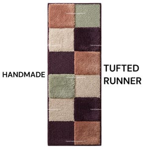 Peut inclure: Un tapis de couloir tufté rectangulaire composé d'une grille de carrés alternés dans des tons vieux rose, vert sauge, crème et prune foncé. Le texte HANDMADE TUFTED RUNNER apparaît sur les côtés de ce revêtement de sol texturé et coloré.