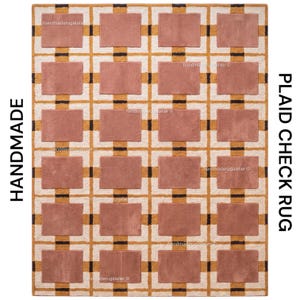 May include: A rectangular rug featuring a repeating grid pattern of dusty rose rectangles framed by white and mustard yellow lines with small black accents. The text HANDMADE and PLAID CHECK RUG is visible on the sides of the rug design.