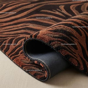 Puede incluir: Una alfombra rectangular con un patrón abstracto similar a una cebra en tonos marrón oscuro y cobre con pelo texturizado. La alfombra está parcialmente doblada para revelar un reverso gris oscuro y un borde acabado con costuras visibles.